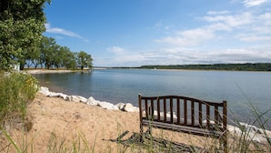 Un banc au bord d’un lac, sur une plage de sable, à l’ombre des arbres.