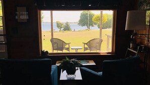 A room with a large window overlooking a grassy area and a body of water. There are two wicker chairs and a small table with a plant on it.