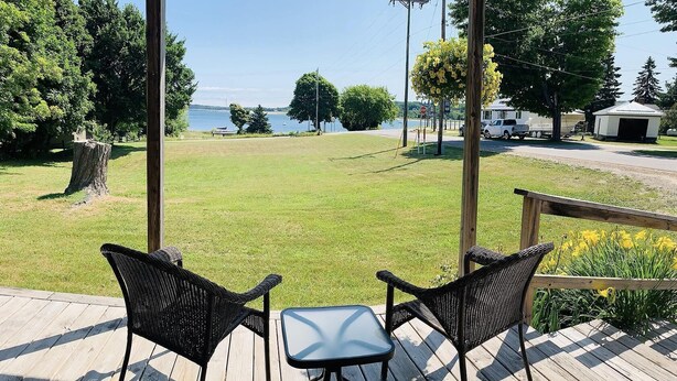 A wooden deck with two wicker chairs and a small table, overlooking a grassy area with a body of water and trees in the background.