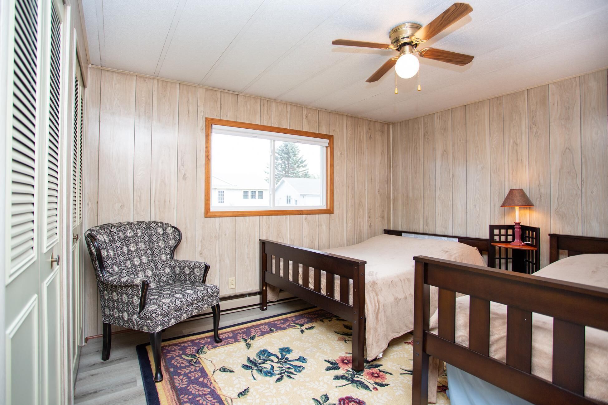 A bedroom with a wooden paneled wall, a single bed with a wooden headboard, a chair with a patterned cushion, a window with a view of a house, and a ceiling fan.