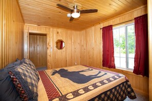 Ein Zimmer aus Holz mit einem Bett, einem Deckenventilator, einem Fenster mit roten Vorhängen und einem Spiegel an der Wand.
