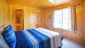 Une pièce aux murs lambrissés, avec un lit, un ventilateur de plafond et une fenêtre avec des rideaux.