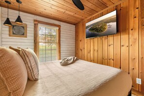 A bedroom with a bed, a window with blinds, a television on the wall, and wooden paneling. - Resort Cabin w/ Firepit, Spacious Deck & Outdoor TV - 2 Miles to SDC (Branson)