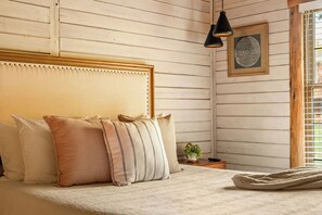 A bedroom with a wooden headboard, a bed with multiple pillows, a bedside table with a plant, and a wall-mounted picture. - Resort Cabin w/ Firepit, Spacious Deck & Outdoor TV - 2 Miles to SDC (Branson)