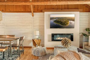 A cozy living room with a wooden ceiling, a dining area, a fireplace, and a large TV mounted on the wall.