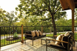 A wooden deck with a sofa and chairs, surrounded by a metal railing, overlooking a road and greenery. - Resort Cabin w/ Firepit, Spacious Deck & Outdoor TV - 2 Miles to SDC (Branson)