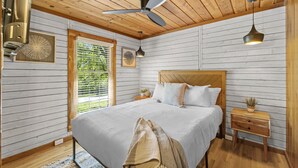 Un dormitorio con cama, techo de madera, ventilador de techo, mesita de noche y una ventana con persianas.