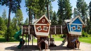 Un parque infantil con dos casitas de madera, un tobogán y un puente de madera.