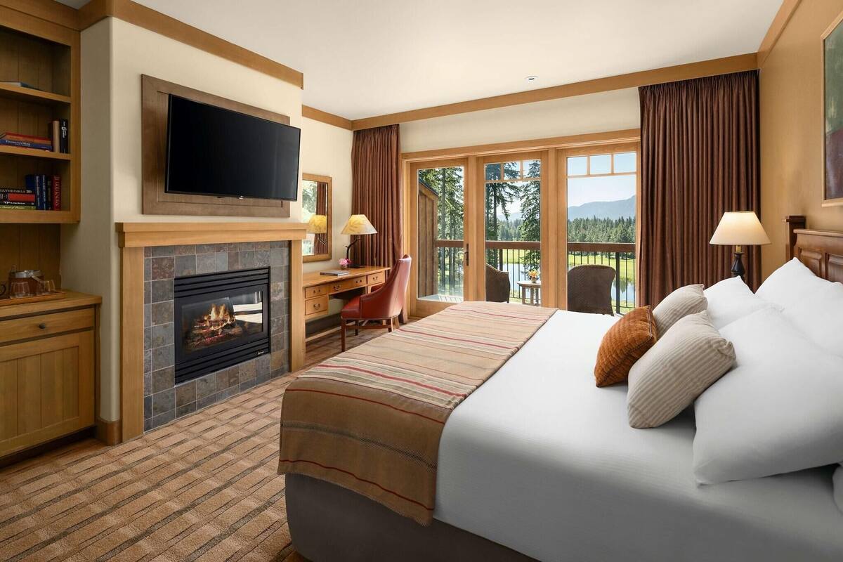 A hotel room with a fireplace, a bed with pillows, a desk with a chair, a television, and a view of the outdoors.