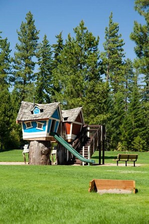 A playground with a slide and a climbing structure, surrounded by a grassy field and trees. - Suncadia Resort (Cle Elum)