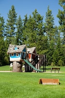 A playground with a slide and a climbing structure, surrounded by a grassy field and trees.