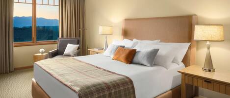 A hotel room with a large bed, two bedside tables, a chair, a window with curtains, and a view of mountains.