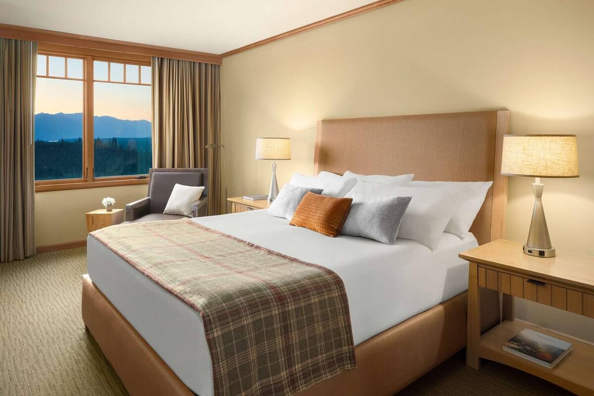 A hotel room with a large bed, two bedside tables, a chair, a window with curtains, and a view of mountains.