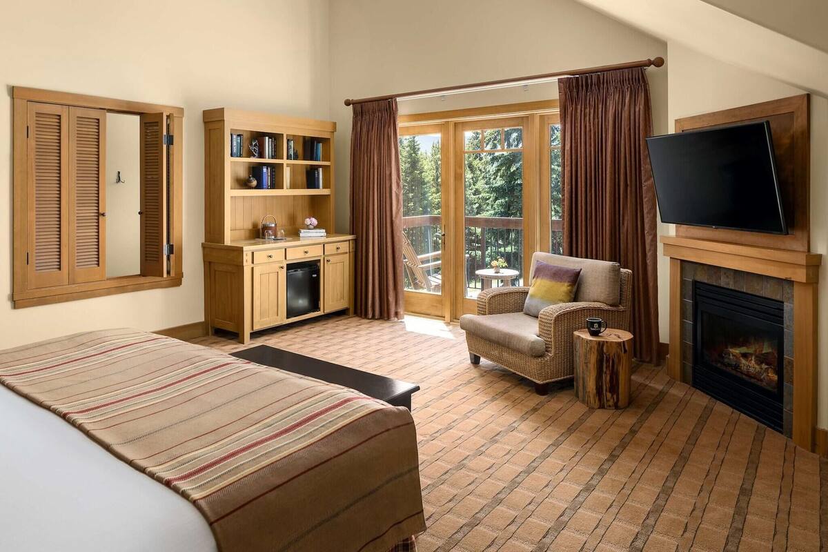 A bedroom with a bed, a television, a fireplace, a bookshelf, and a view of the outdoors.