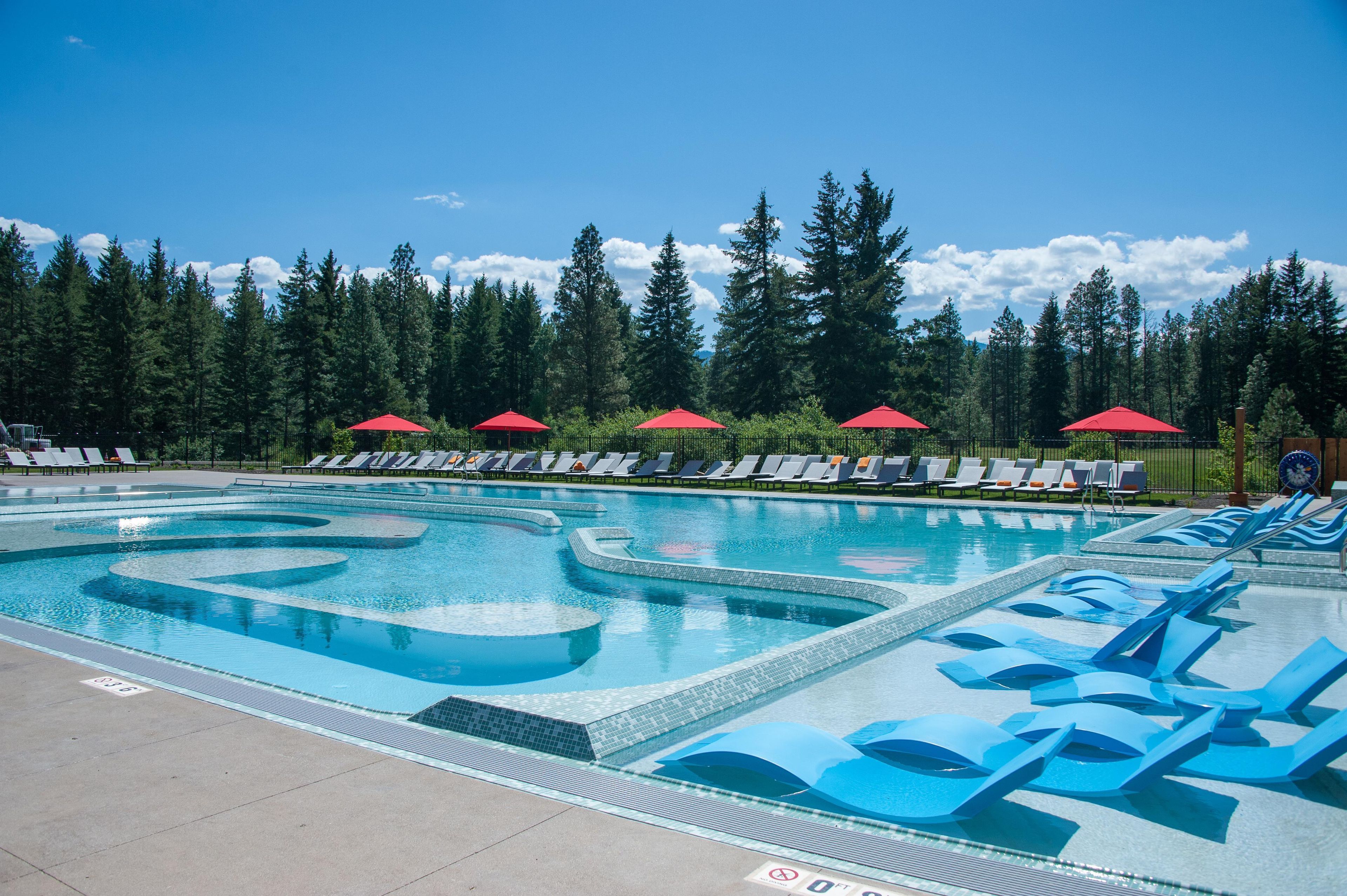 A swimming pool with blue and white patterns, surrounded by lounge chairs and red umbrellas, with a forested background.