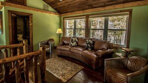A cozy living room with a leather sofa, a wooden coffee table, and a view of trees through large windows. - Spectacular Views, Private Estate , Hot Tub, Close to Everything (Intervale)