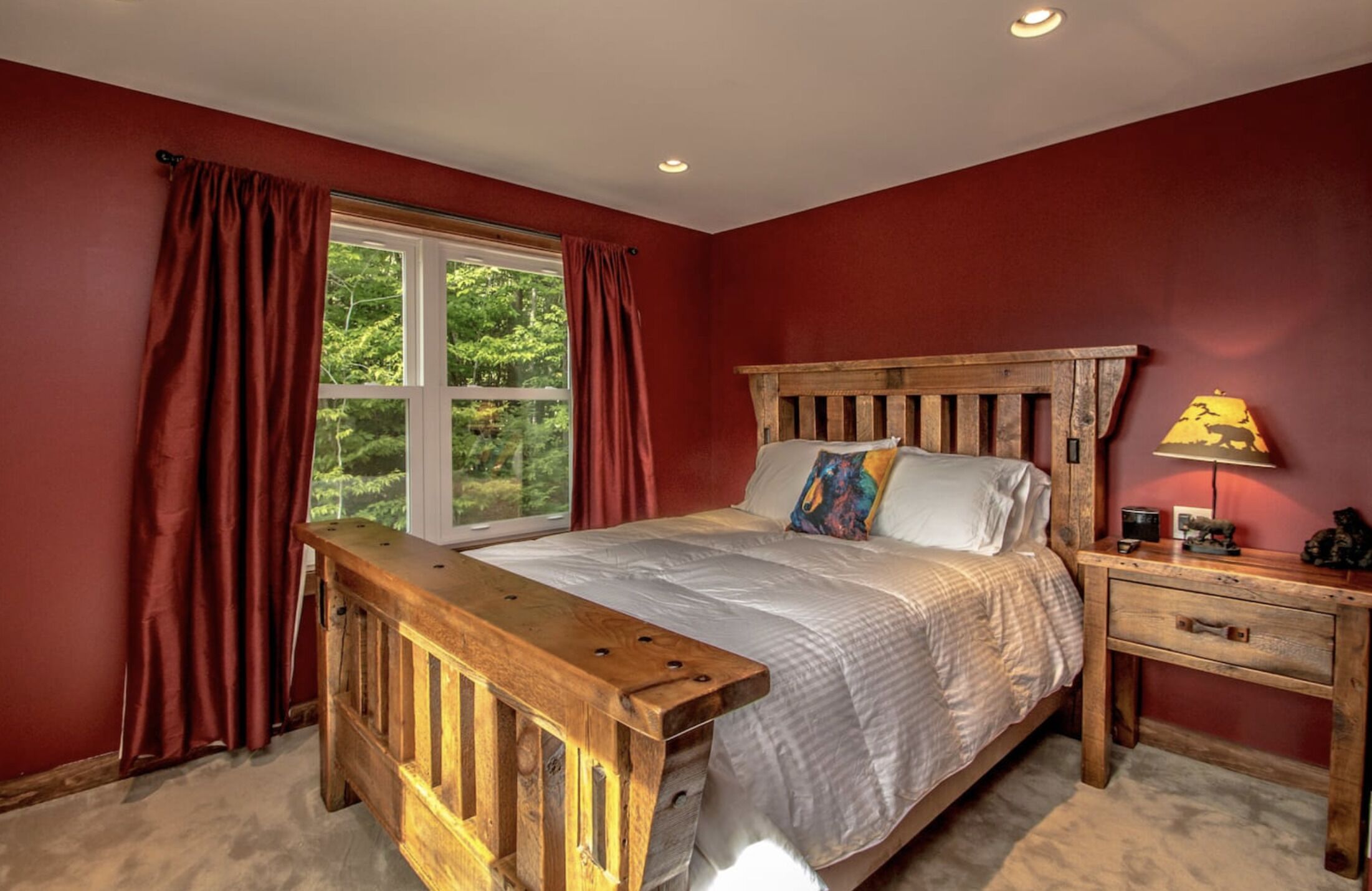 A bedroom with a wooden bed, red walls, a window with curtains, a bedside table, and a lamp.