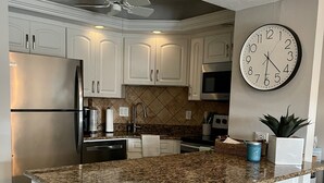 A kitchen with white cabinets, a granite countertop, a stainless steel refrigerator, a microwave, a clock, and a ceiling fan. - Walk out the back door to #1 beaches in Naples. Renovated with new Furnishings. (Naples)