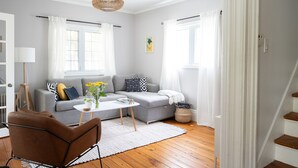 A modern living room with a grey sofa, a brown armchair, a white coffee table, and a wicker pendant light. - Steps to Main Street, Picton w Sandbanks Pass, Bbq and Large Private Back Yard (Picton)