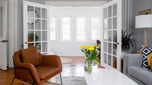 A living room with a brown leather chair, a grey sofa, a white coffee table, and a vase with yellow flowers. - Steps to Main Street, Picton w Sandbanks Pass, Bbq and Large Private Back Yard (Picton)