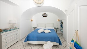 Ein Schlafzimmer mit weißer Rundbogenzelle, einem Bett mit blauer Tagesdecke, weißen Kissen, einer Glastafel und einer Pendelleuchte.