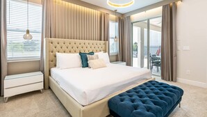 A modern bedroom with a tufted headboard, a large bed with white linens, a blue ottoman, and a view of the outdoors through a sliding glass door. - 11BR Grand Villa with Epic Game Room Near Disney (Kissimmee)