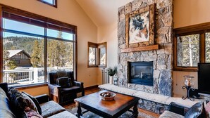 Ein gemütliches Wohnzimmer mit Kamin aus Stein, großen Fenstern und Blick auf eine Holzhütte.