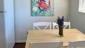 A dining area with a wooden table, white chairs, a ceiling fan, and a wall-mounted painting of a crab. - Dungeness Bay Bungalow- The Ultimate Couples Retreat (Sequim)
