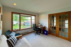 A room with a large window, a desk with a chair, a couch with striped pillows, and a blue trash bin.