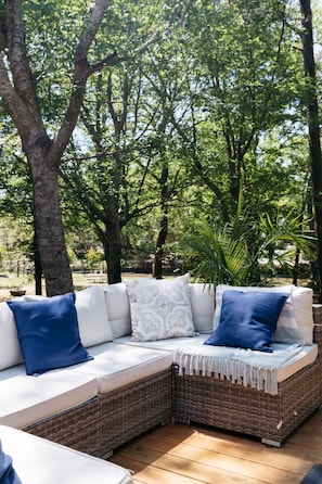 An outdoor wicker sofa with white cushions and blue pillows, set on a wooden deck surrounded by trees.