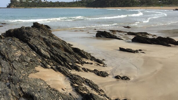 A rocky beach with waves, a sandy shore, and a forested hill in the background.