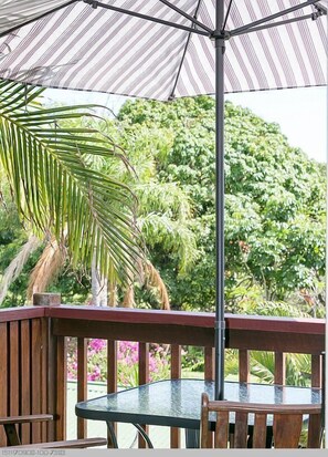An outdoor patio with a glass table and wooden chairs, surrounded by a wooden railing and a striped umbrella. - Boat Harbour Studio Apartments and Villas (Hervey Bay)