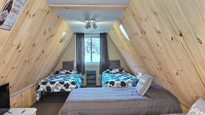 Ein gemütliches Dachzimmer mit Holzwänden, zwei Betten mit floralem Bettzeug, einem Deckenventilator und einem Fenster mit Vorhängen.
