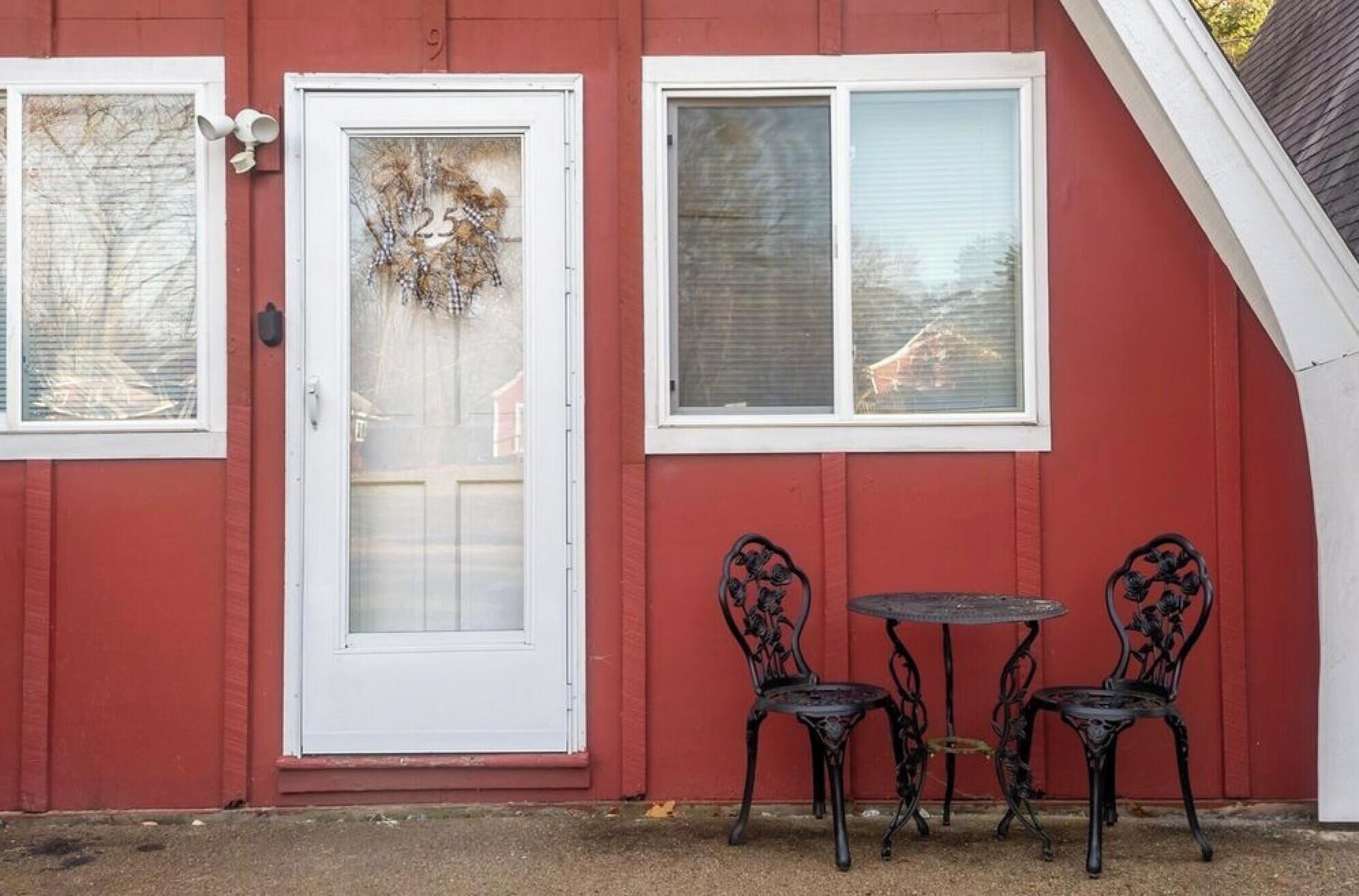 Un bâtiment rouge avec une porte blanche, deux fenêtres et deux chaises en métal noir, accompagnées d’une petite table.