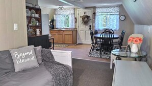 Ein gemütliches Wohnzimmer mit grauem Sofa, Couchtisch und Essbereich mit Glastisch und Stühlen. Im Hintergrund ist eine Küche mit Holzausbau und einem Fenster erkennbar.