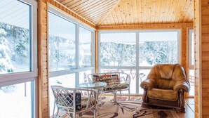 Ein gemütliches Zimmer mit Holzwänden und schrägen Decken, mit einem großen Fenster und Blick auf schneebedeckte Bäume. Ein Ledersessel und ein Glastisch mit Stühlen sind ebenfalls vorhanden.