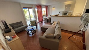 Un salon comprenant un canapé, une table basse et un téléviseur. Il y a également un coin cuisine avec un four à micro-ondes et un ventilateur.