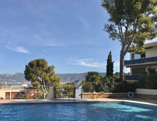 A swimming pool with a view of mountains in the background, surrounded by trees and a building with balconies.