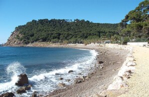 A beach with pebbles, a rocky shoreline, and a forested hill in the background.