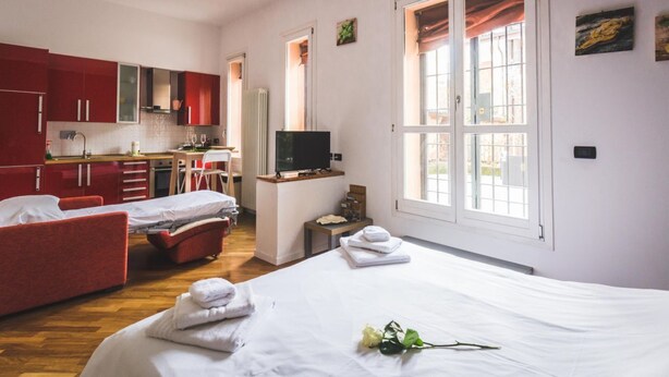 A hotel room with a bed, two red armchairs, a small table with a TV, a kitchen area, and large windows. - GetTheKey San Felice Suite 1 (Bologna)