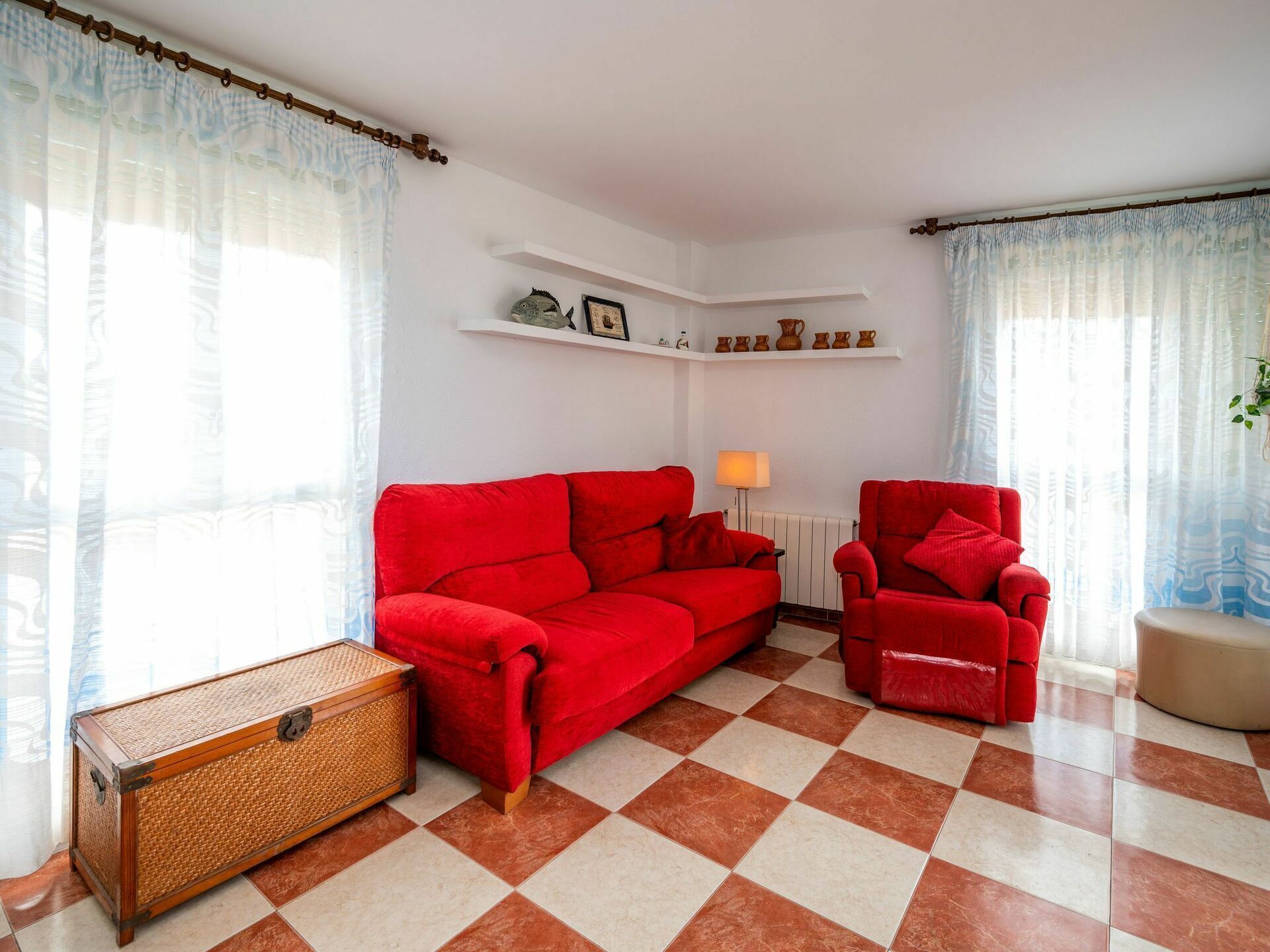 A living room with a red sofa, a red armchair, a wicker chest of drawers, and a tiled floor.