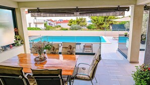 Una terrazza esterna con tavolo e sedie in legno, una piscina sullo sfondo e una zona coperta con vista sul quartiere.