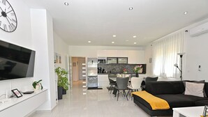 Un soggiorno moderno con una TV a schermo piatto, un divano nero, un tavolo da pranzo e una cucina sullo sfondo.