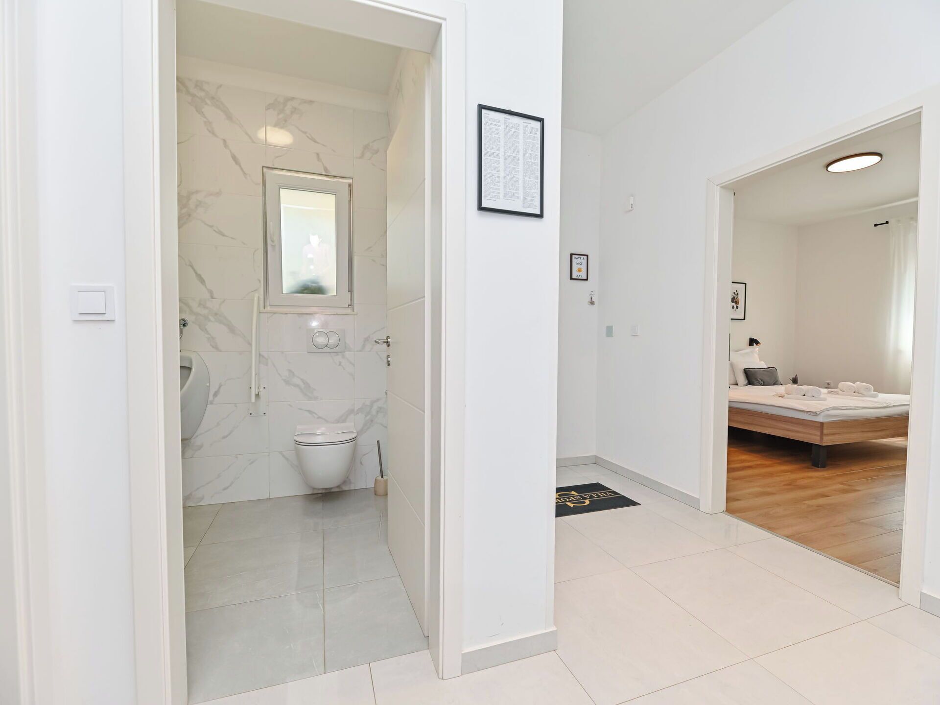 A modern bathroom with a marble wall, a toilet, and a shower. There is a framed sign on the wall and a door leading to a bedroom with a bed and a nightstand.