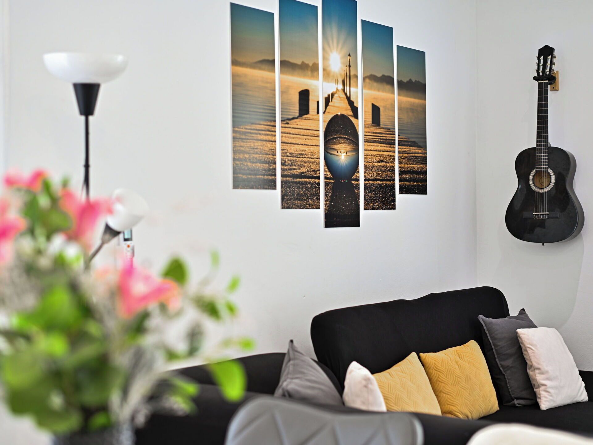 Een woonkamer met een zwarte bank, een gitaar en een muur met een zonsondergangschilderij.