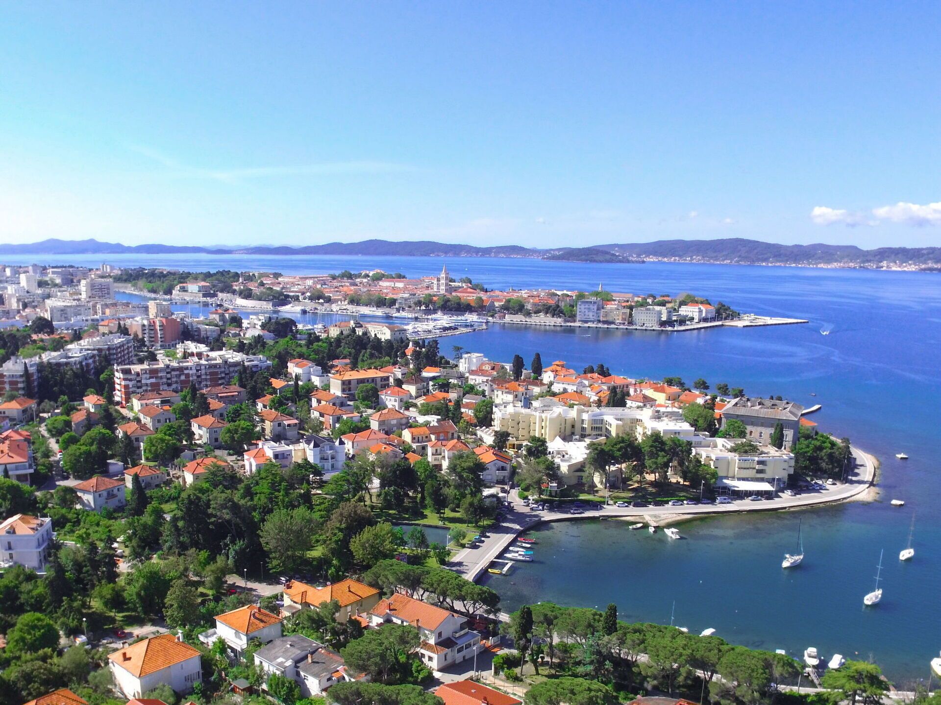 A coastal cityscape with a marina, buildings with red-tiled roofs, and a clear blue sea.