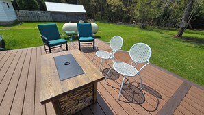 A wooden deck with a table and chairs, surrounded by a grassy yard and trees. - Drummond Island Hotel & Vacation Homes - Island Gem (Drummond)