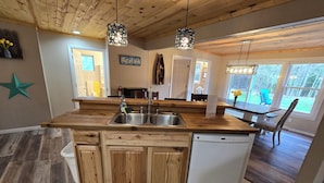 A kitchen with wooden cabinets, a dishwasher, and a large island with a sink. There are pendant lights, a window, and a view of the outdoors. - Drummond Island Hotel & Vacation Homes - Island Gem (Drummond)