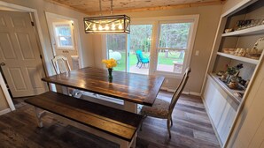A wooden dining table with benches, a hanging light fixture, and a view of a backyard through large windows. - Drummond Island Hotel & Vacation Homes - Island Gem (Drummond)