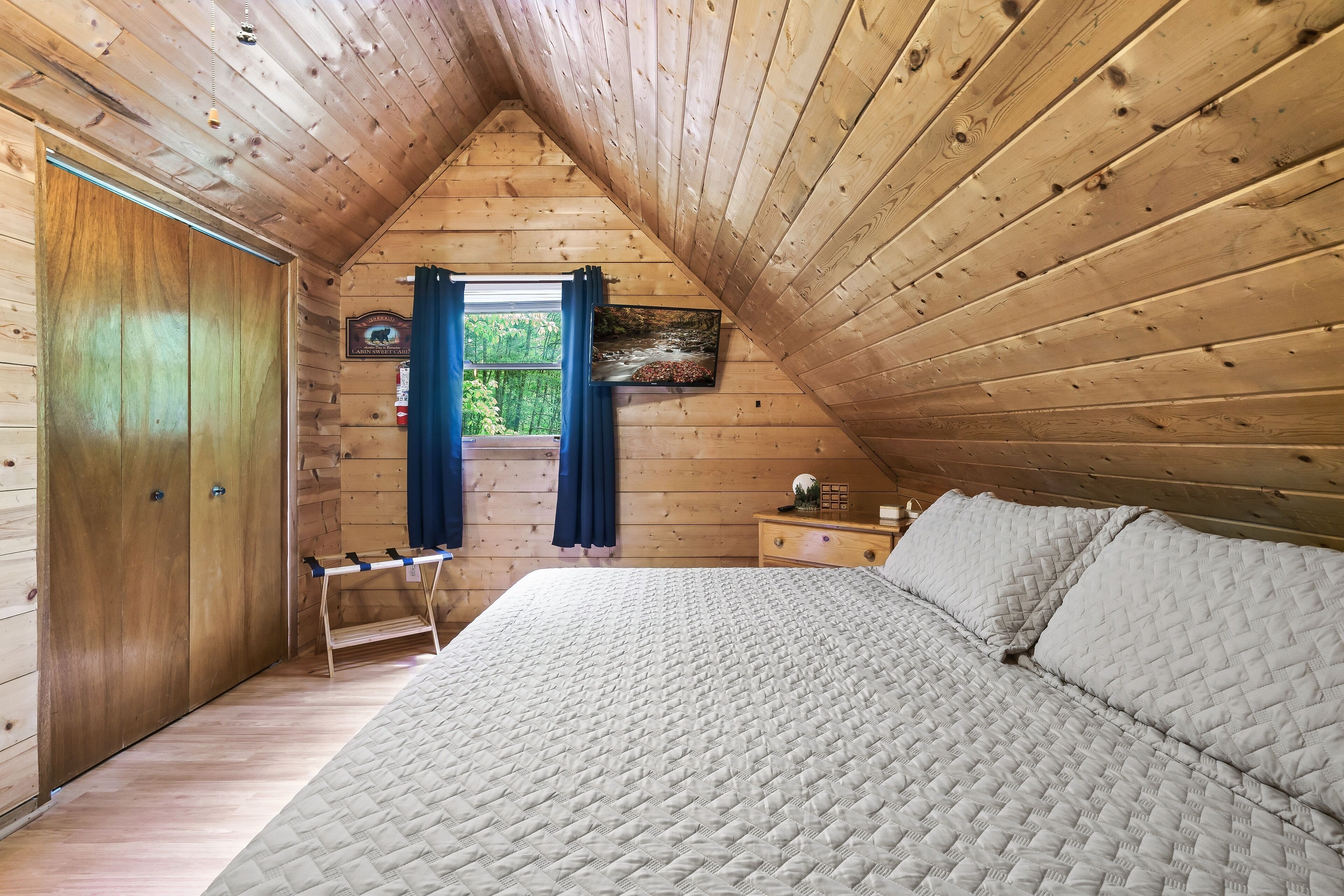 Un dormitorio con techo de madera, una cama con cubrecama acolchado, una mesita de noche, una ventana con cortinas y una puerta.